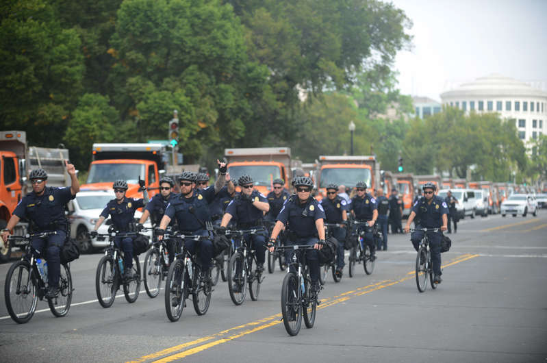 a group of people riding bikes down a street: Long lines of trucks, used as security barricades, and plenty of law enforcement gather along 3rd Street where demonstrators gather for the Justice for J6 rally near the U.S. Capitol. (Photo by Astrid Riecken For The Washington Post)